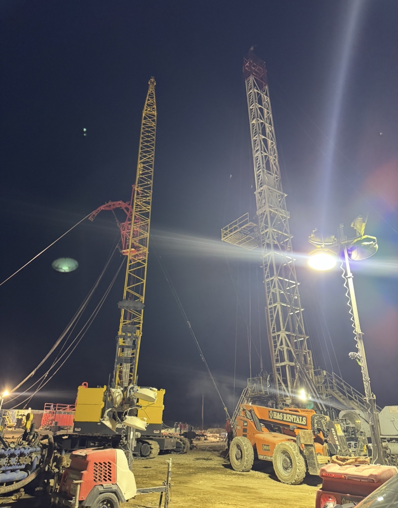 Crane supporting coiled tubing beside a rig mast under lights at night